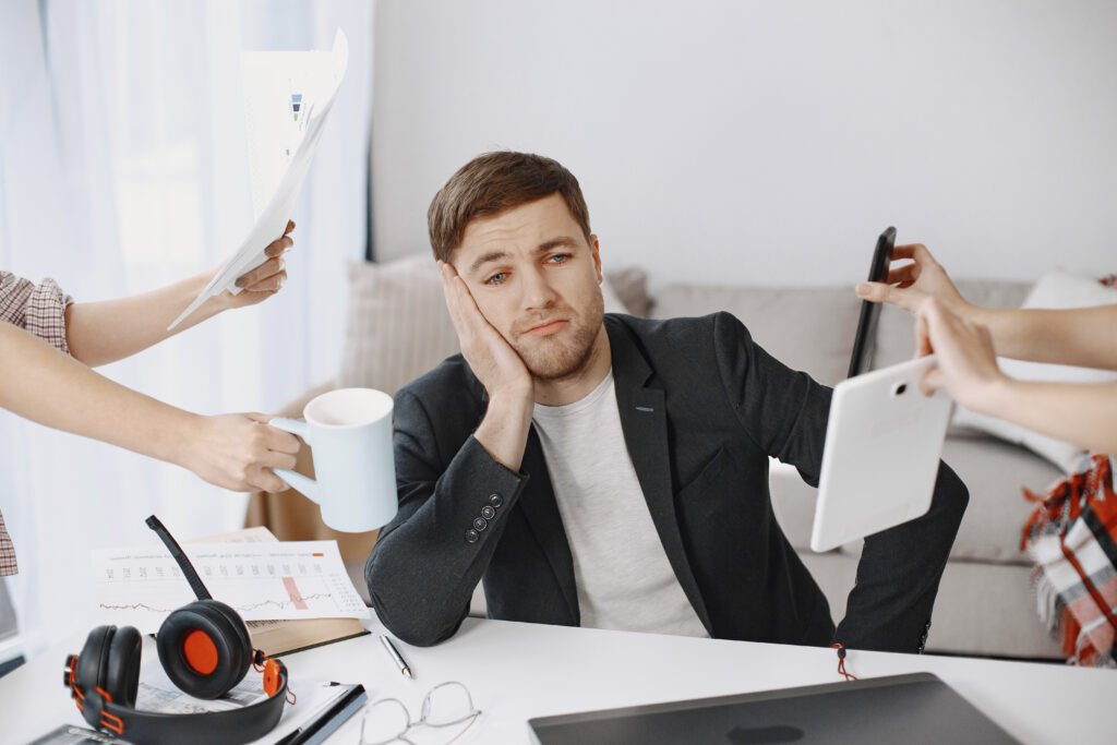 Man sitting at a desk looking tired and overwhelmed while several hands around him hold papers, a mug, a phone, and a tablet, symbolizing work pressure and multitasking stress.